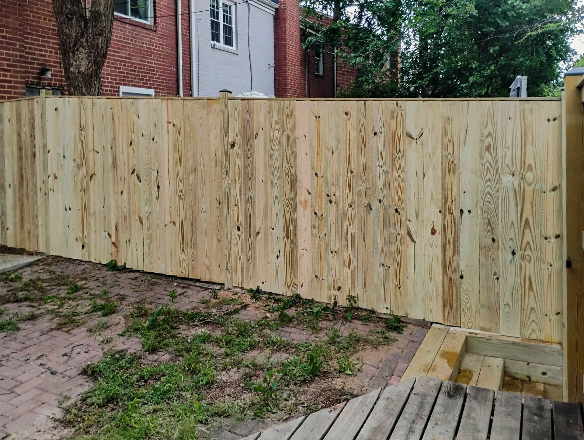 Wooden fence in a backyard; brick building visible in background, some overgrown grass in foreground.