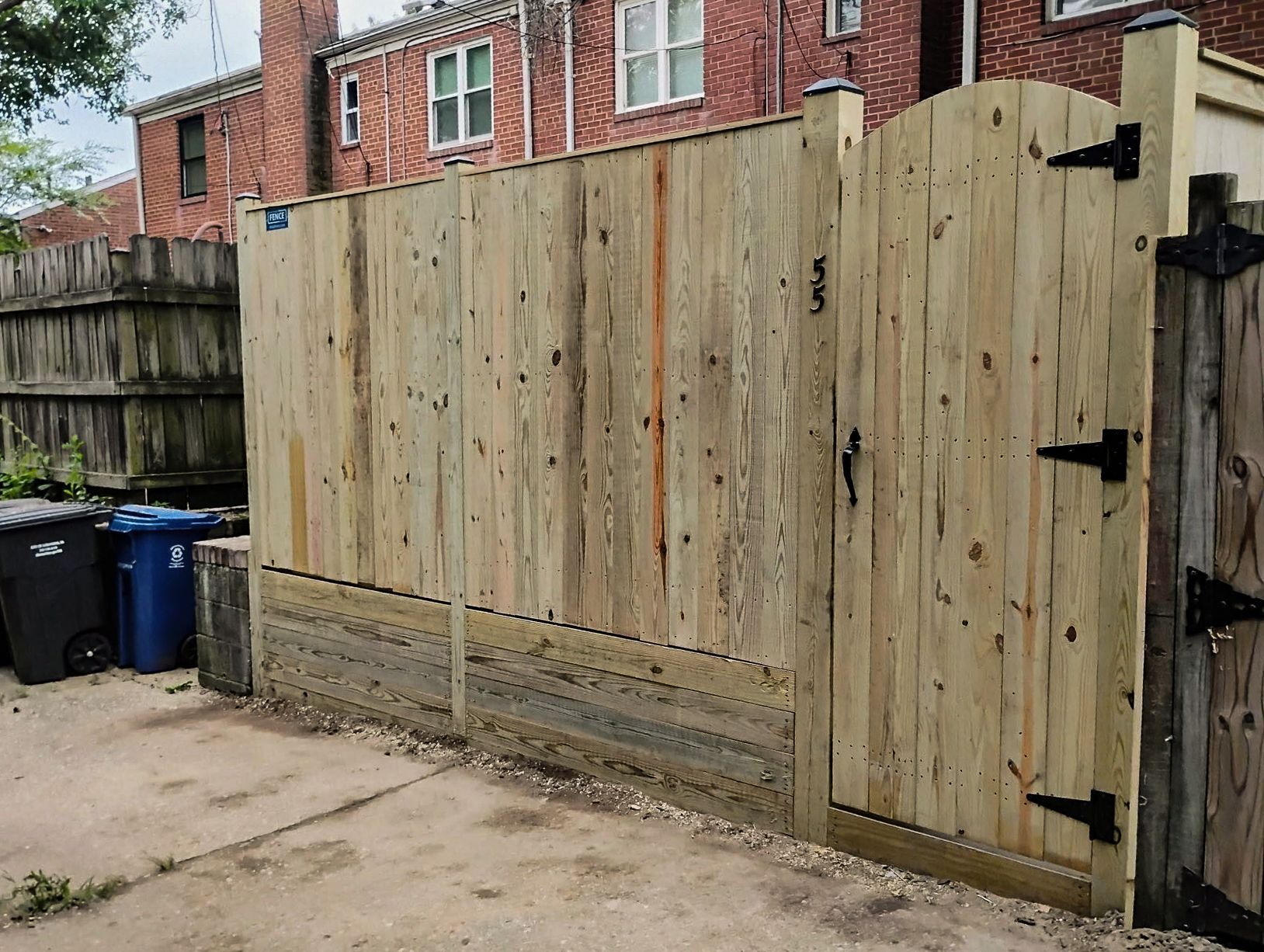 Wooden fence with gate, light brown, with trash cans on the left and a building in the background.