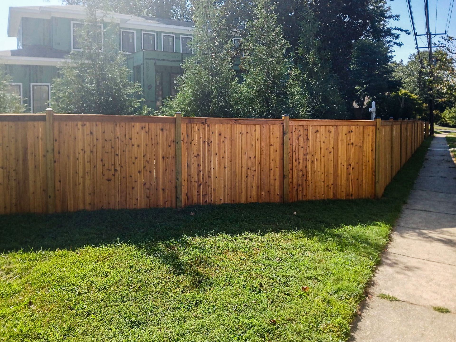 Wooden privacy fence along a green lawn and sidewalk, with a two-story house in the background.