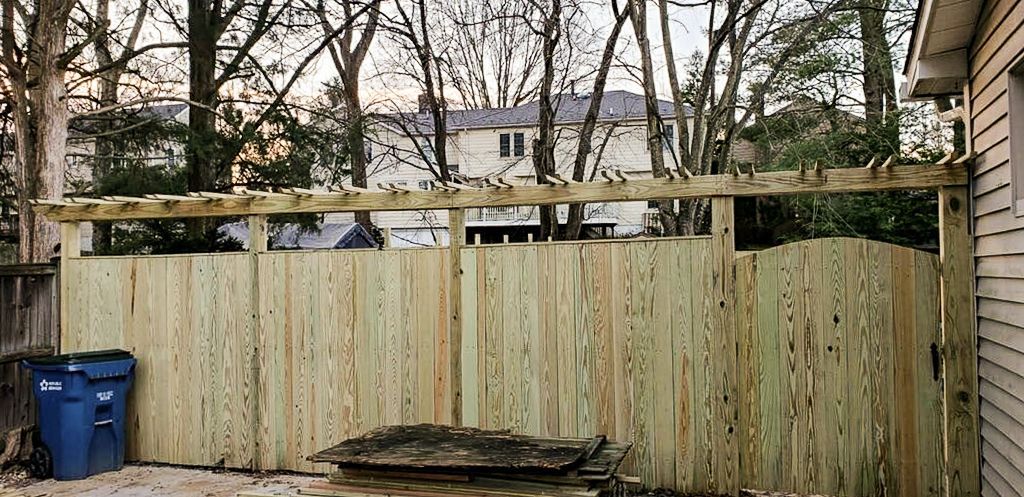 Wooden fence with a pergola-like structure on top, behind trees and a house. Blue trash bin on the left.