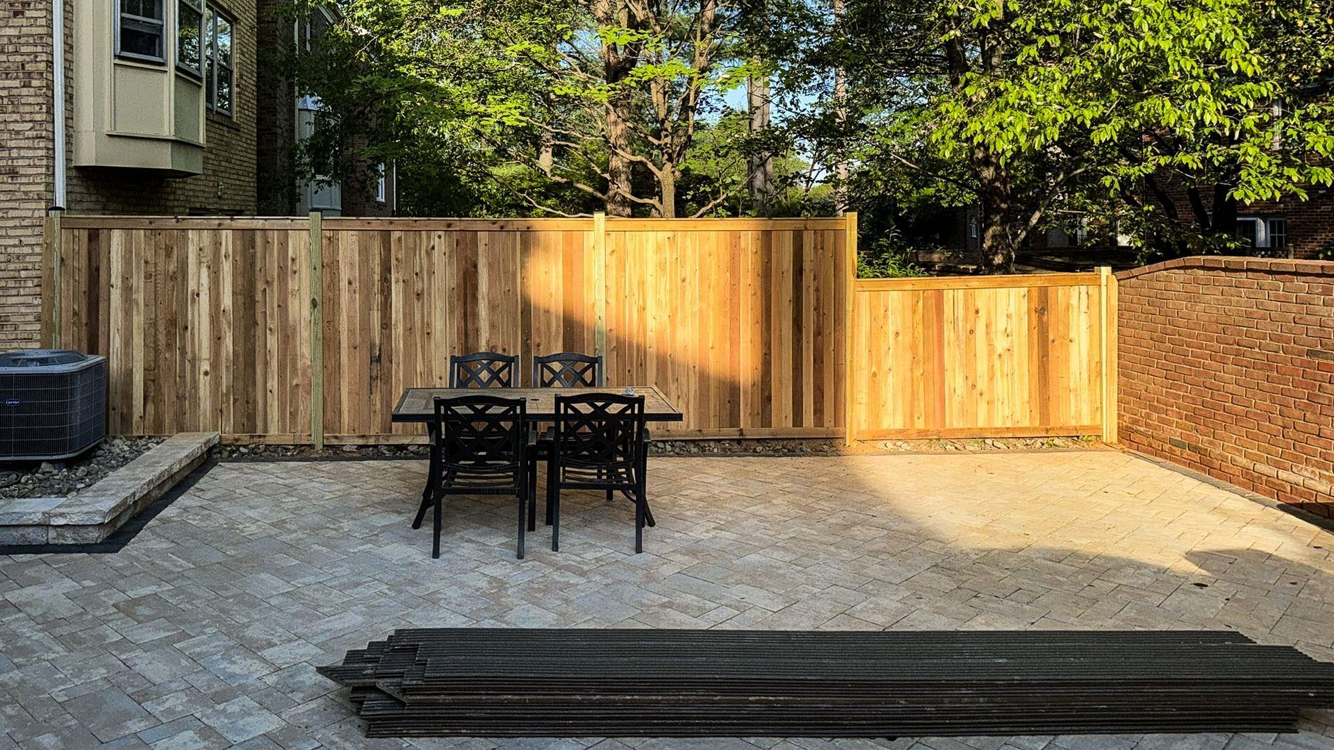 Patio with a wooden fence, dining table, and trees in the background. Sunlight shines on the area.