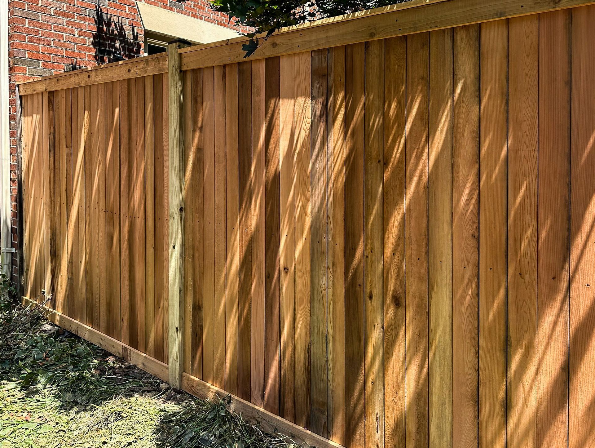 Wooden privacy fence casting shadows, against a brick wall, grass in foreground.