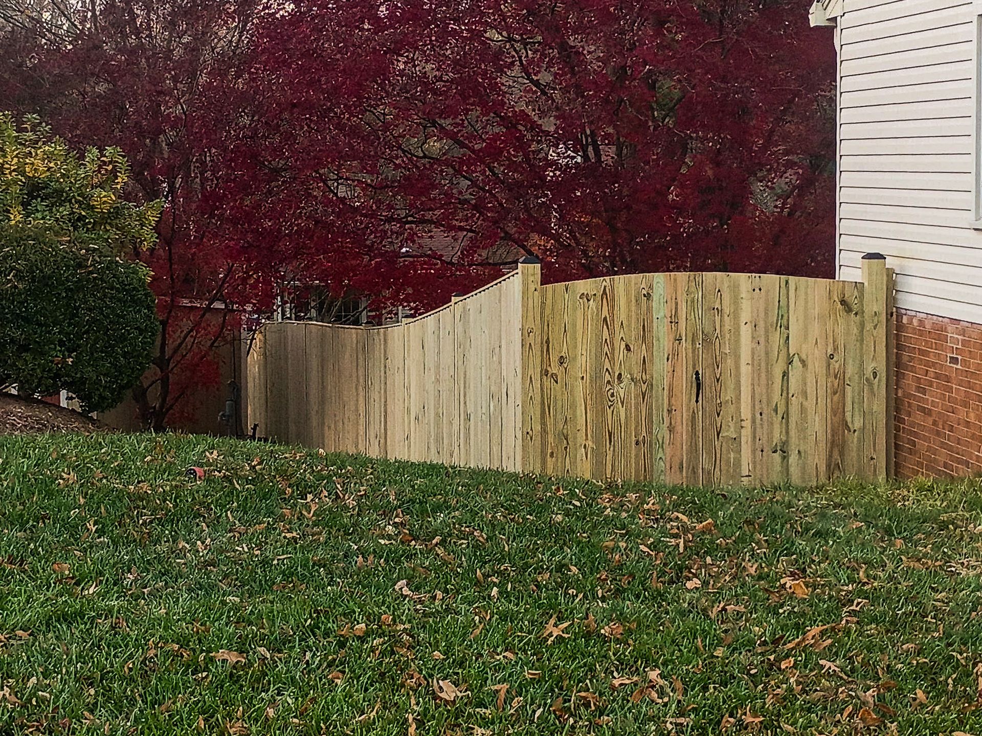 Wooden fence with an arch shape, in front of red-leafed tree, on a grassy hill near a house.