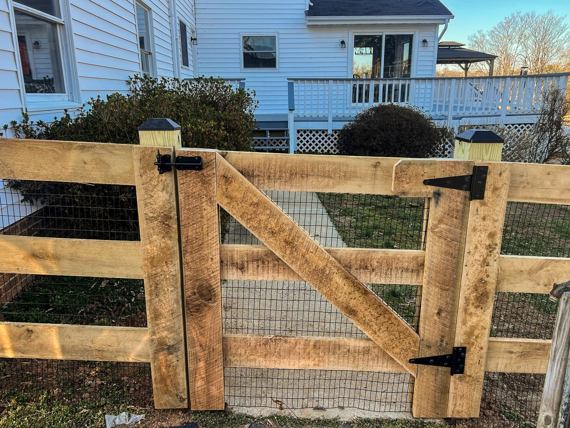 Wooden gate in a tan fence, leading to a brick path and white house with deck.