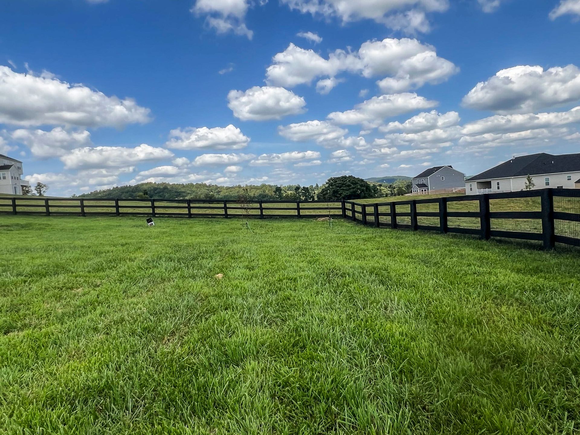 Grassy field enclosed by black fence, with trees and homes in background under a partly cloudy sky.