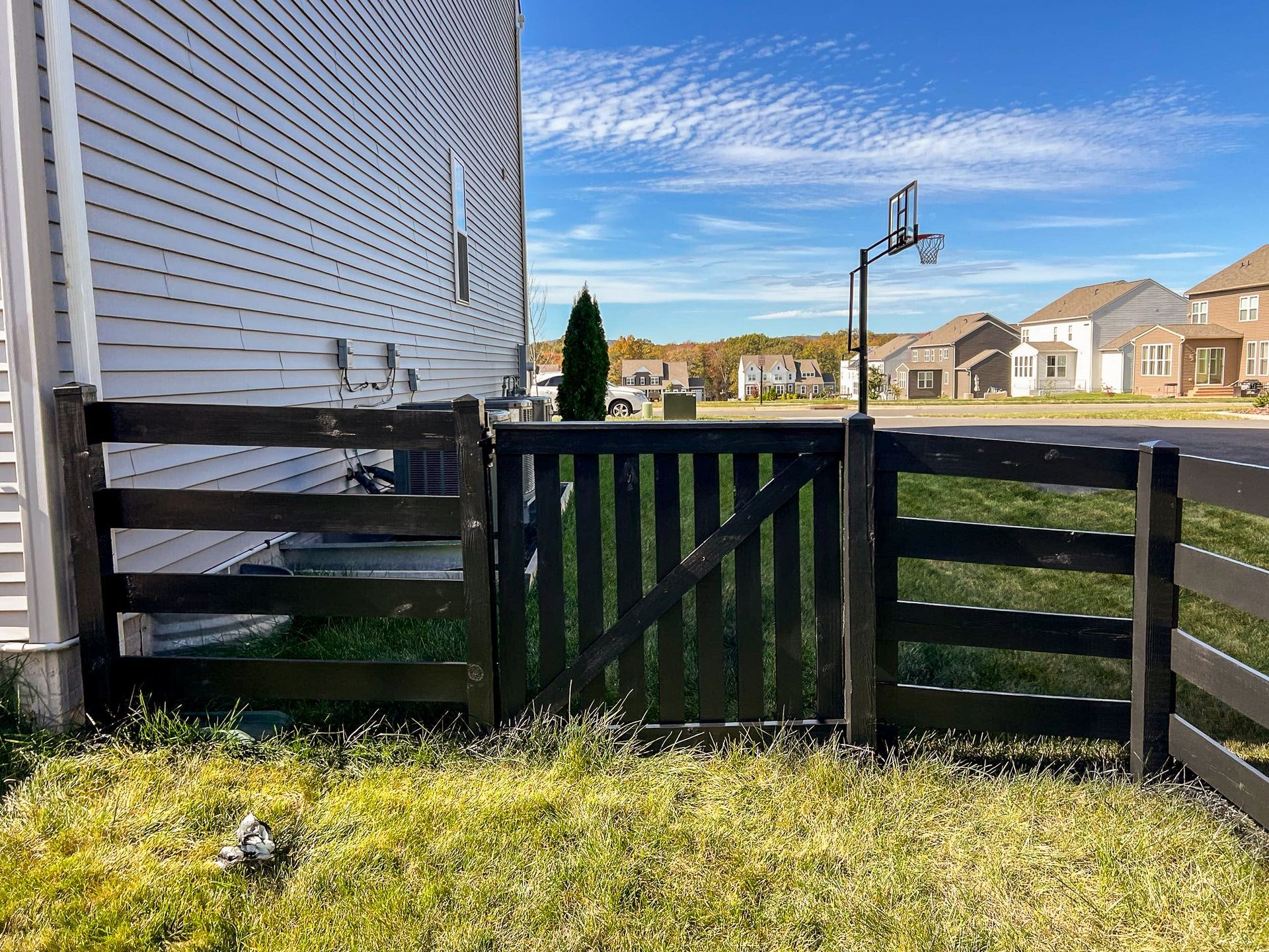 Black wooden fence with gate in front of a house, basketball hoop visible in the distance.