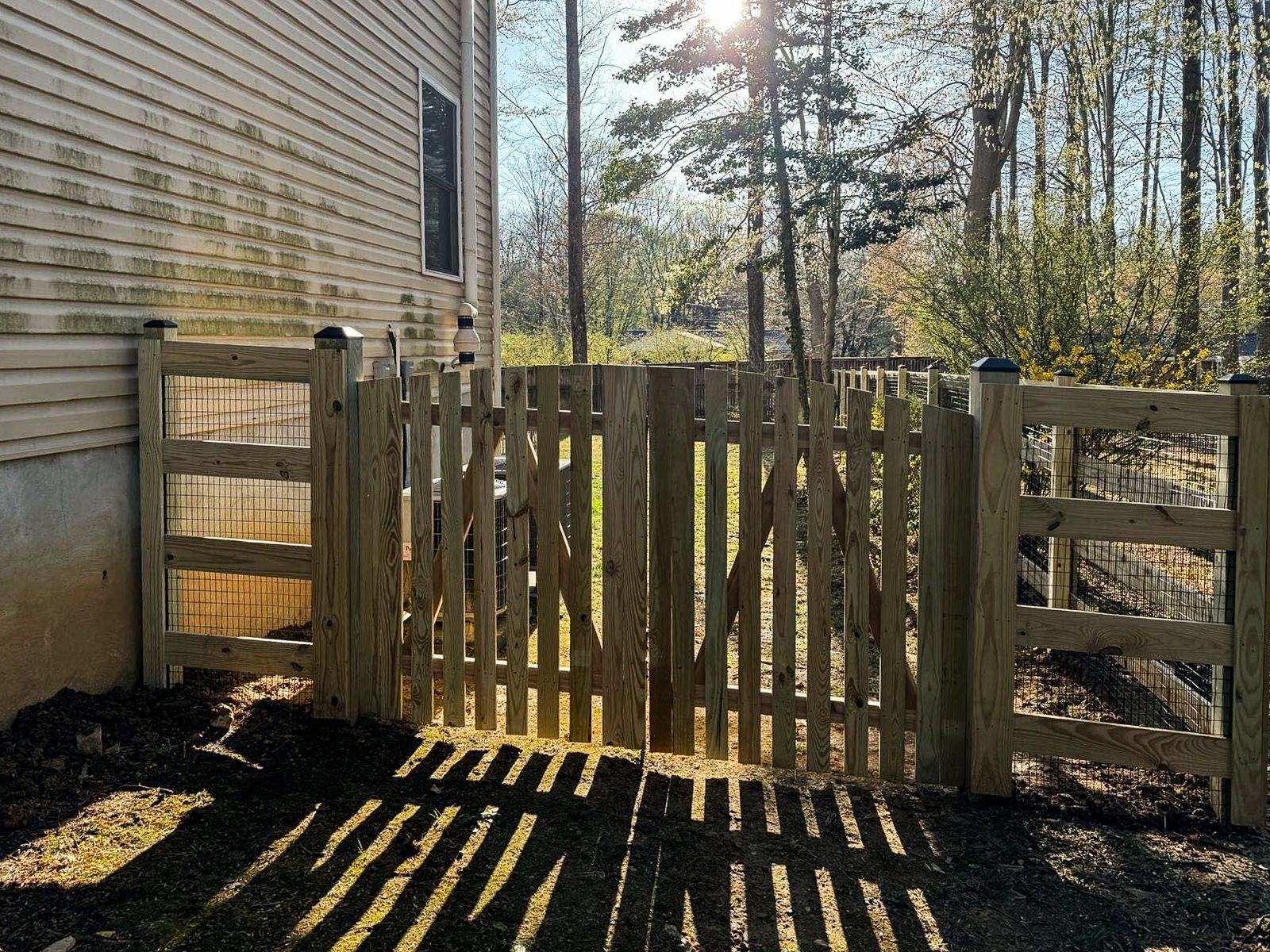 Wooden gate in a backyard, with a house to the left and trees in the background, casting long shadows.