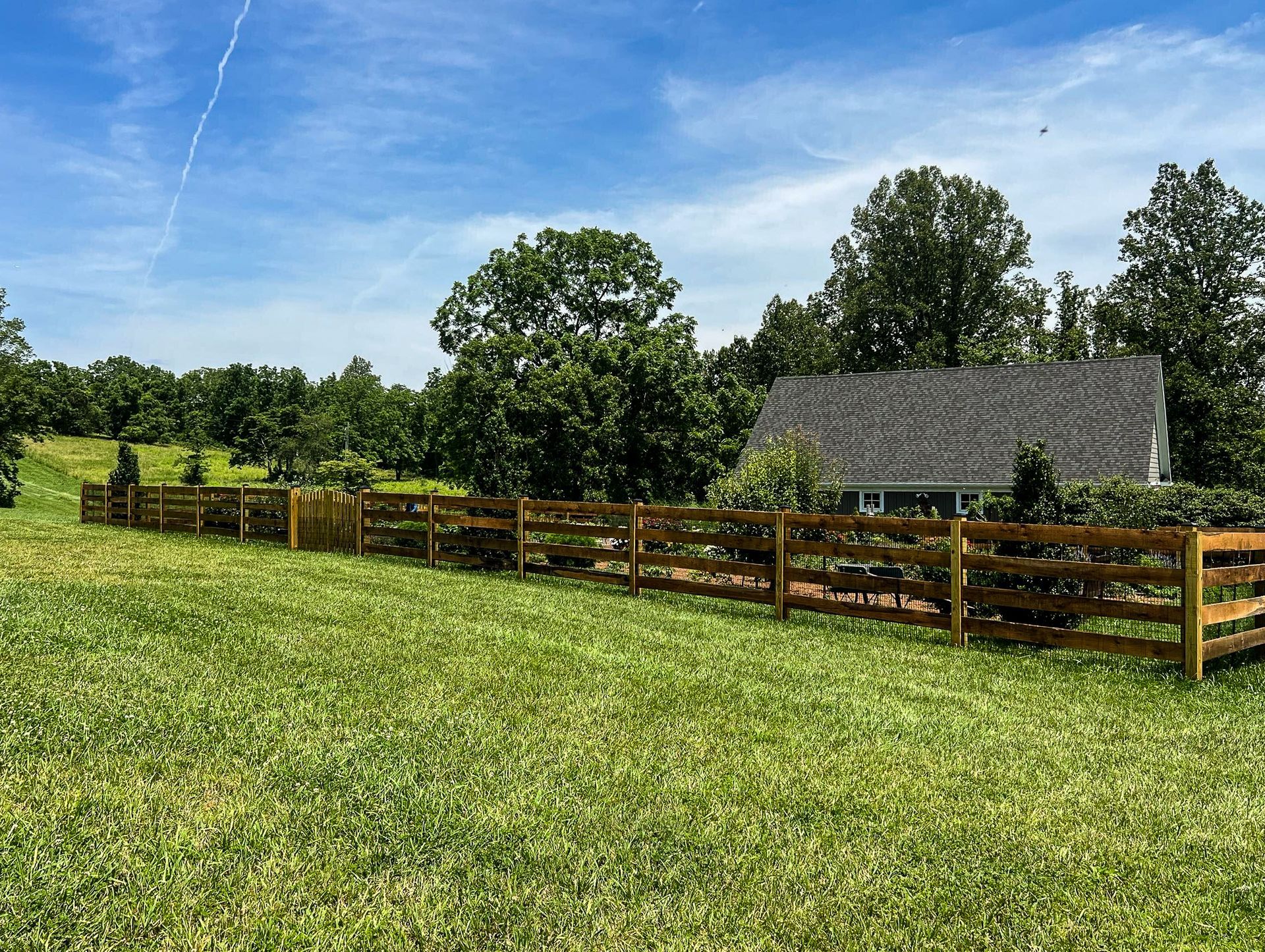 Wooden fence surrounds a grassy field with a house and trees in the background under a blue sky.