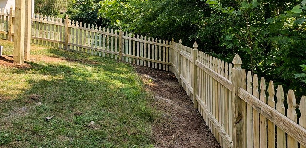 Wooden picket fence along a grassy yard and dirt path, with trees in the background.