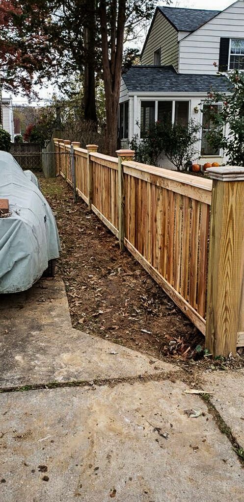 Wooden fence alongside a driveway, a covered car on the left, and a house in the background.