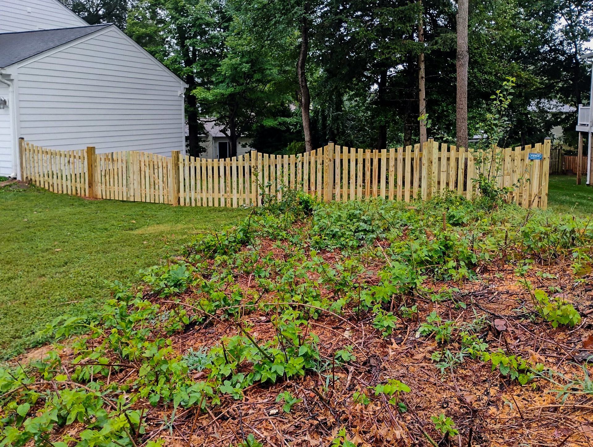 Wooden picket fence curving across a grassy yard, with a house visible in the background and hillside in the foreground.