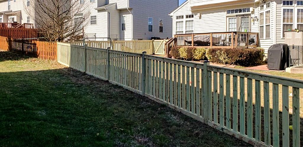 A wooden picket fence on a grassy hill in front of houses with a backyard deck.