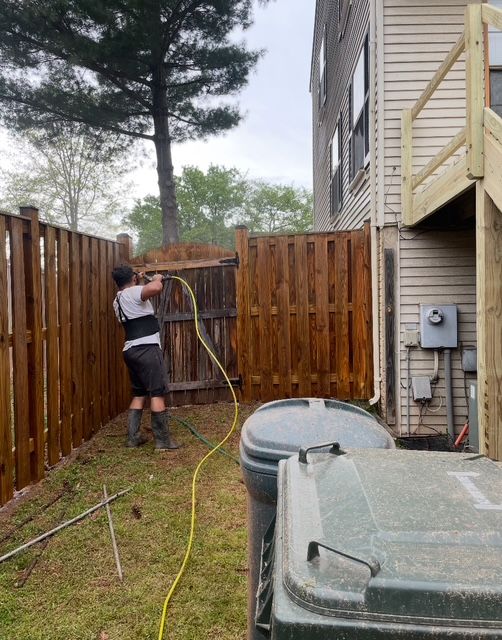 Person power washing a wooden fence in a backyard. The fence is brown. A trash can is in the foreground.