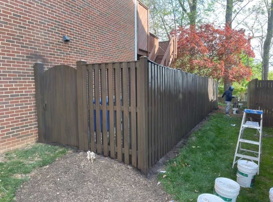 A wooden fence painted brown, next to a brick building. Person painting the fence outside.
