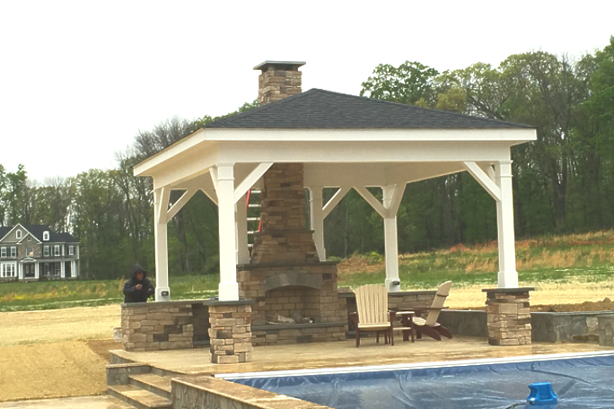 Poolside gazebo with stone fireplace and chimney. Black roof, white trim. Two chairs visible.