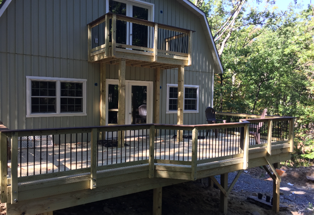 Wooden deck attached to a light green house with upper balcony. Black railings and surrounding trees are visible.