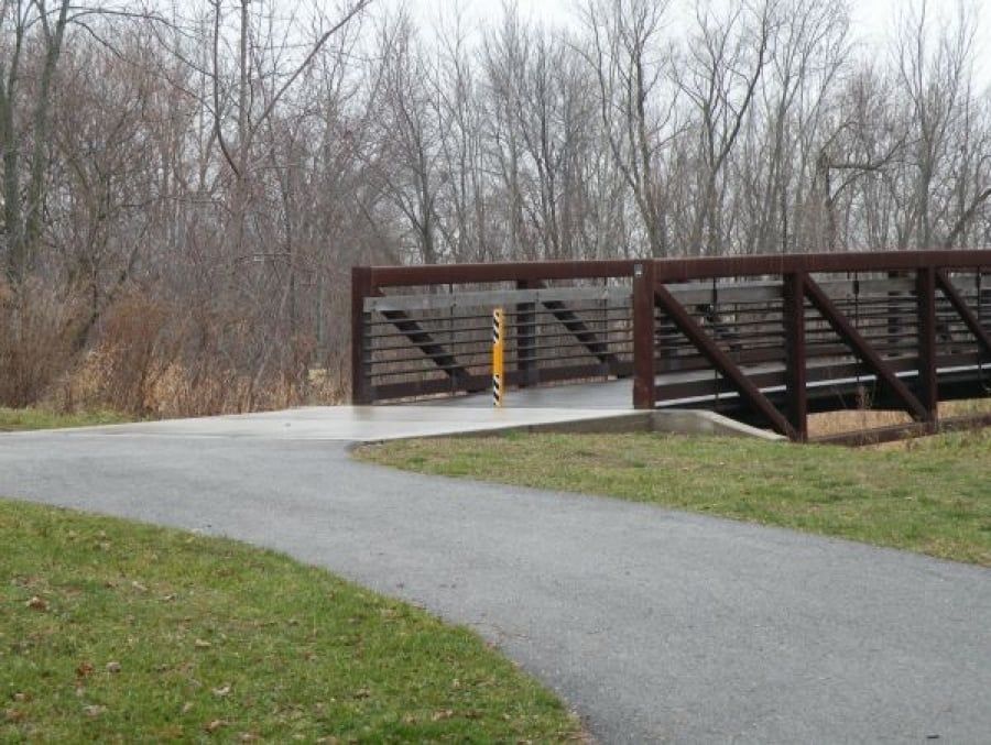 Paved path leading to a brown wooden bridge over a creek in a park. Bare trees in the background.
