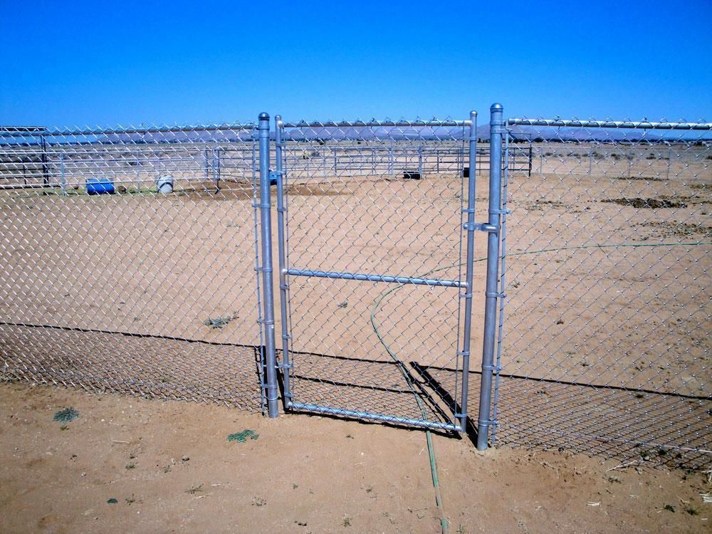 Chain link fence with open gate in a desert setting under a bright blue sky.