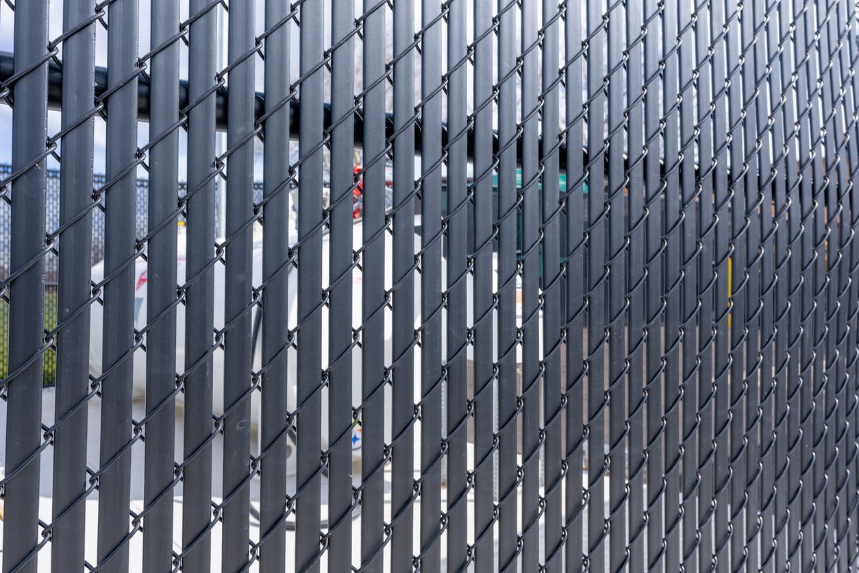Gray chain-link fence with vertical privacy slats, partially obscuring a blurred background.