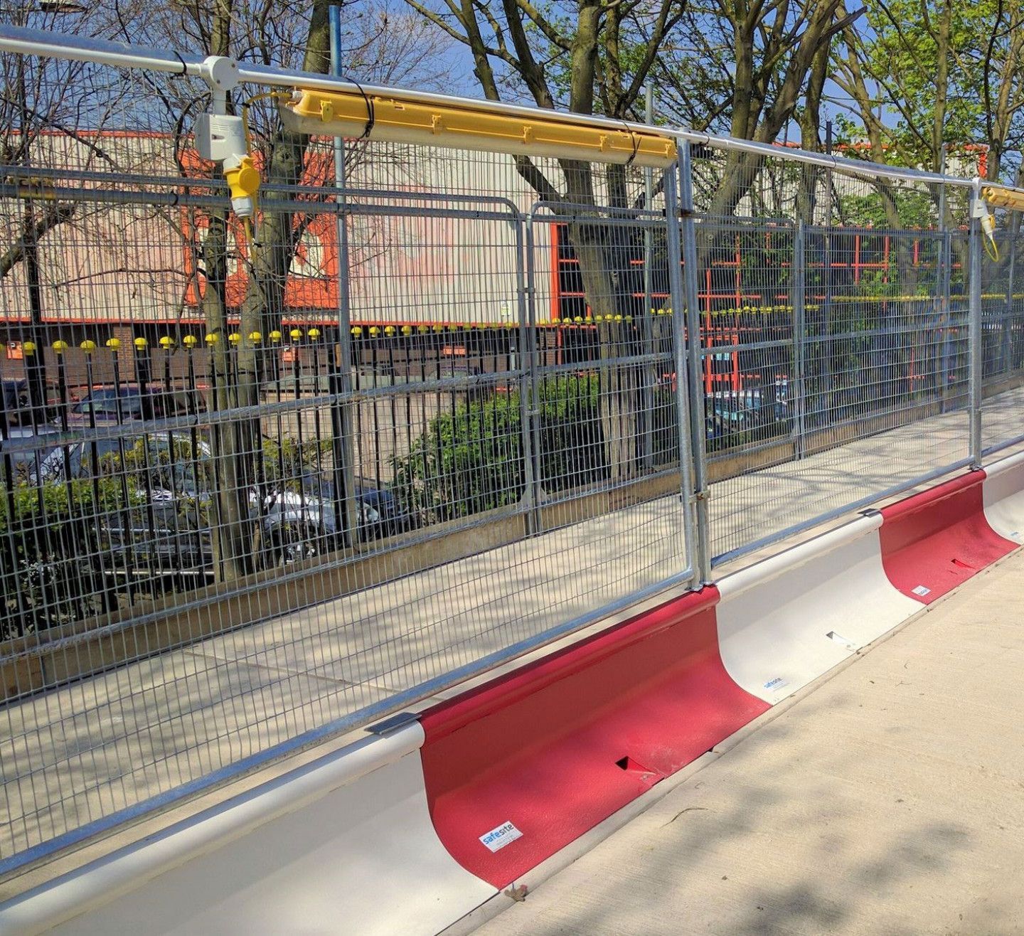 Metal fence with red and white barrier alongside a sidewalk, trees, and buildings in the background.