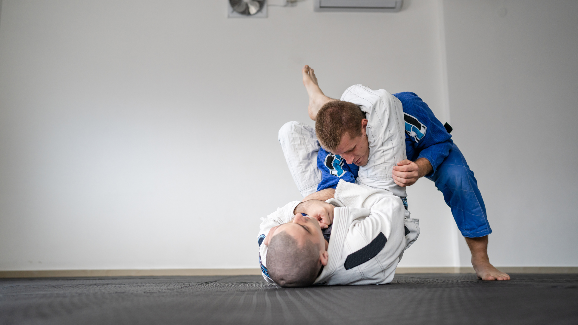 Two men in blue and white gis grappling on a mat indoors. One is on top, leg hooked, applying pressure.