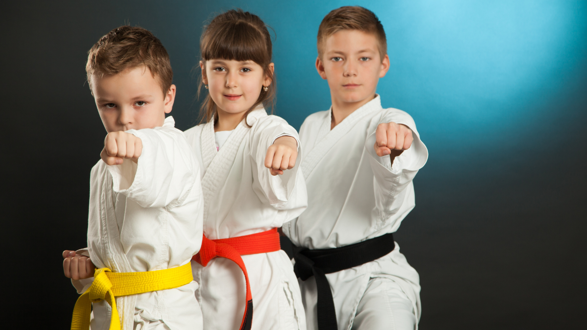Three children in karate uniforms: yellow, red, and black belts, in a karate stance with fists clenched.