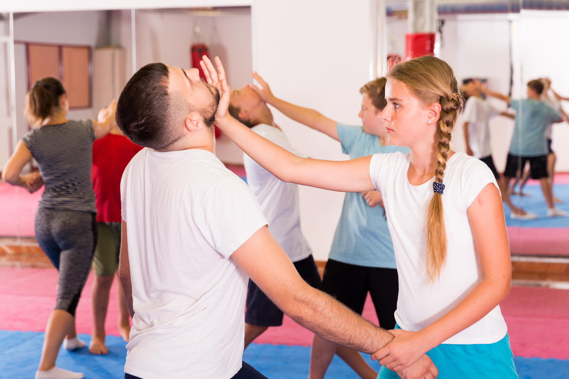 Martial arts class: A woman practices a face strike on a man in a gym. Other people are in the background.
