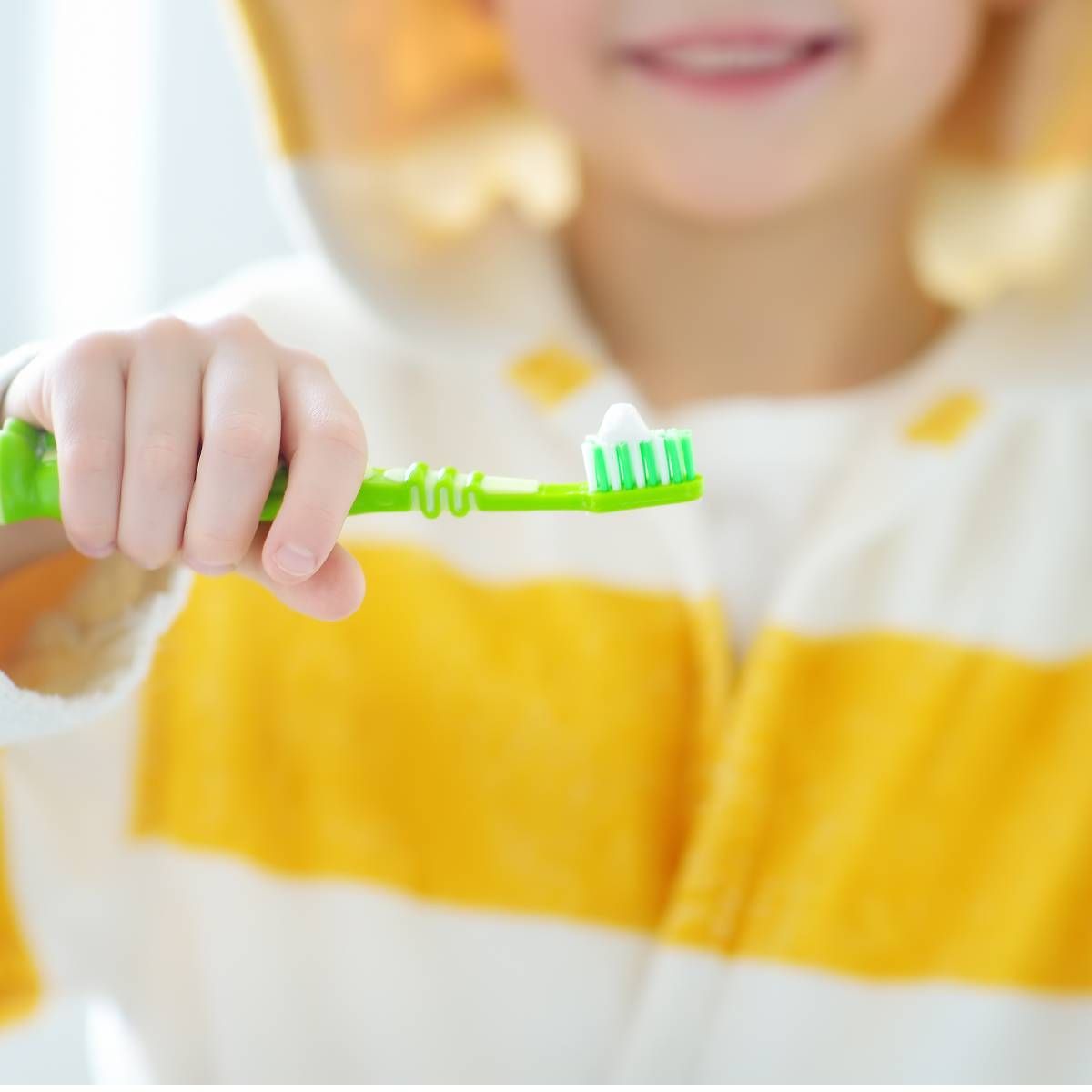 Child holding a green toothbrush with toothpaste, smiling. Wearing yellow and white striped robe.