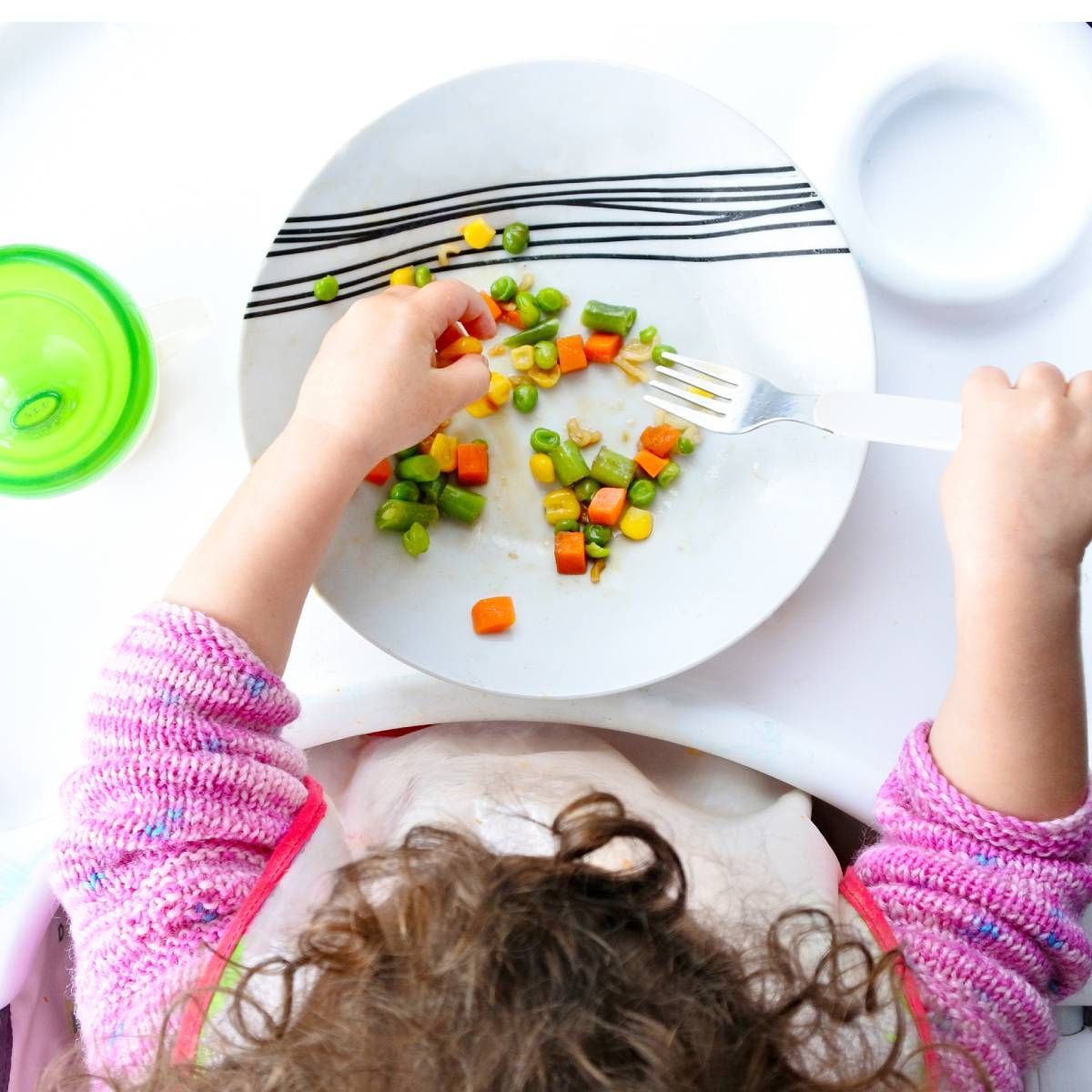 Child in pink sweater reaches for vegetables on a plate, fork in hand.