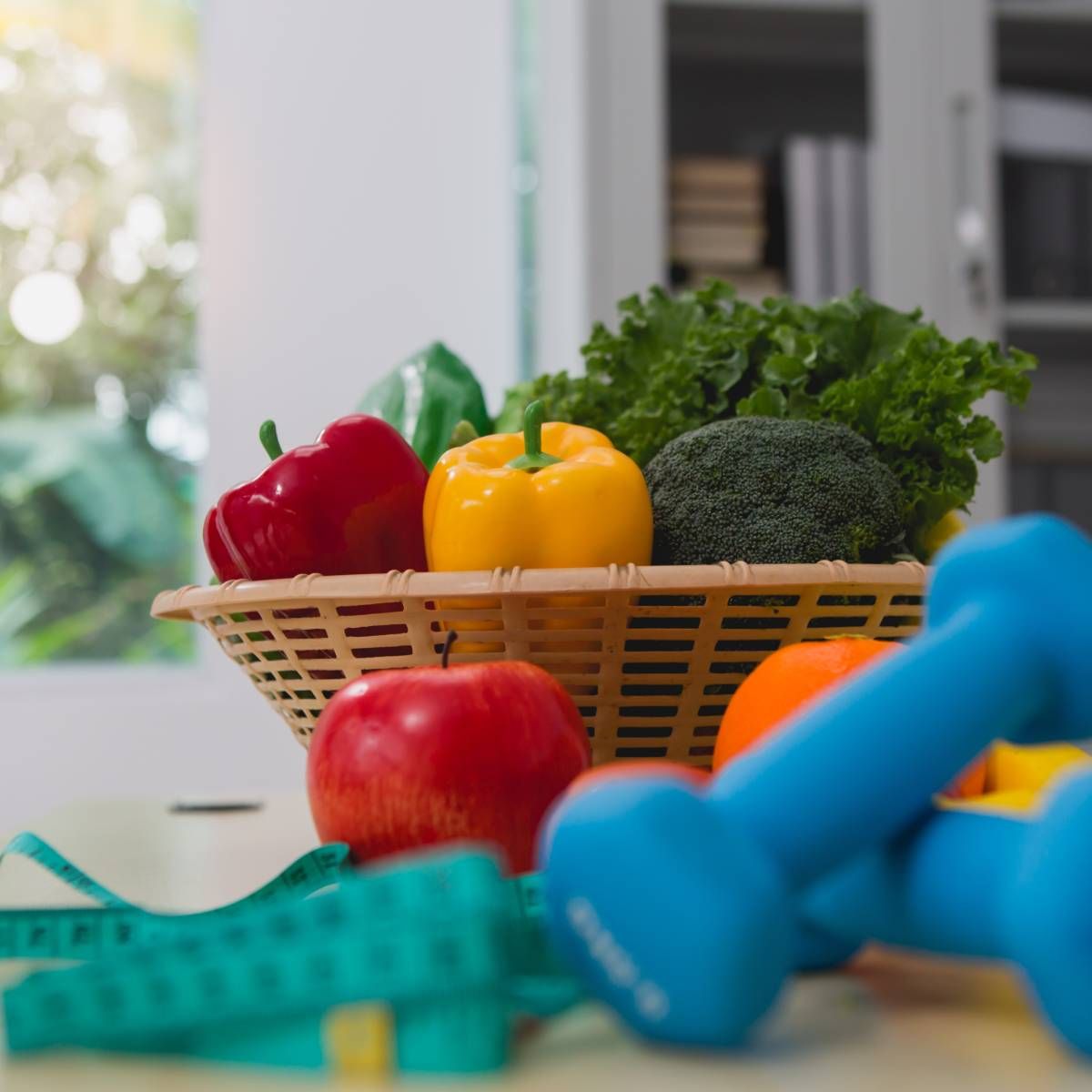Basket of colorful vegetables and fruit with dumbbells and a measuring tape, suggesting healthy eating and exercise.