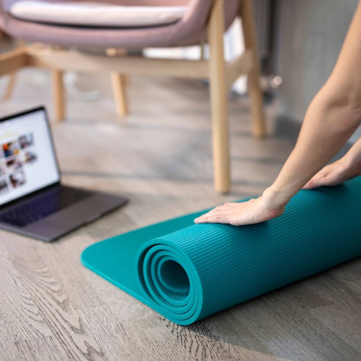 Person rolling out a teal yoga mat on a wooden floor with a laptop open in the background.