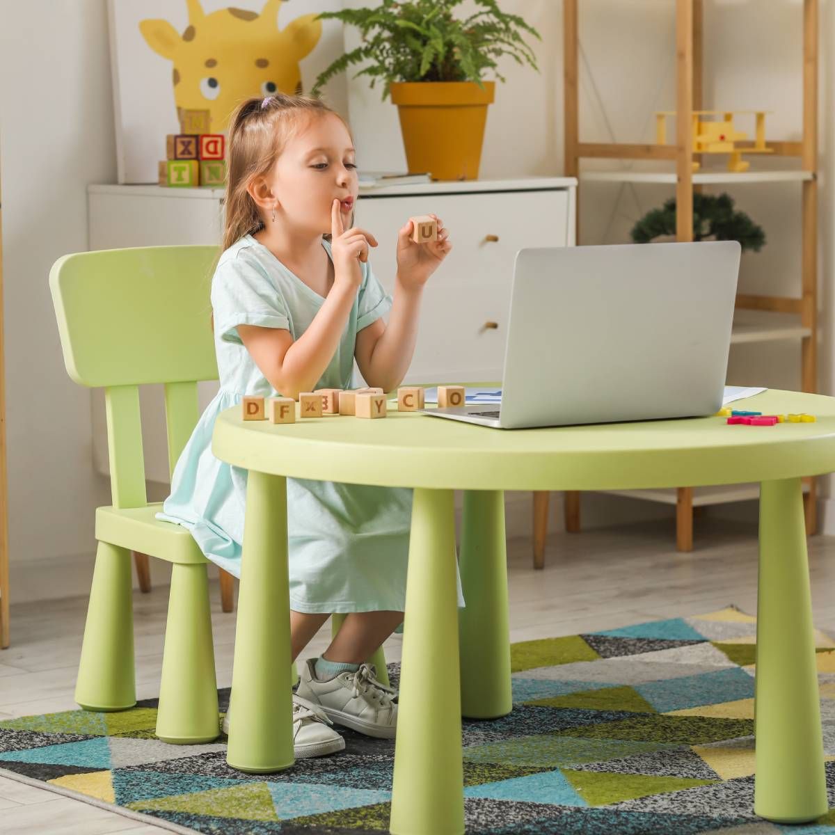 Girl at a table, fingers to lips, using laptop and letter blocks in a light-green room.