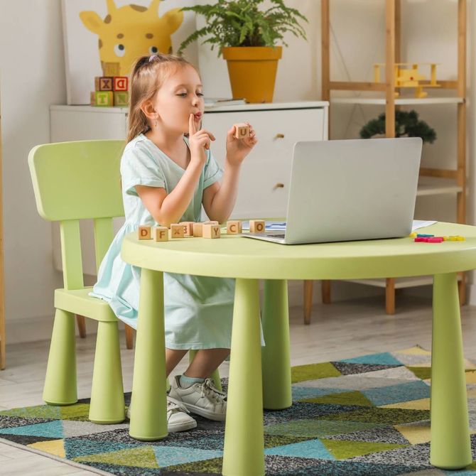 Girl at a table, fingers to lips, using laptop and letter blocks in a light-green room.