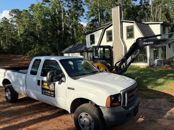 White utility truck parked beside a house with a black backhoe in the yard.