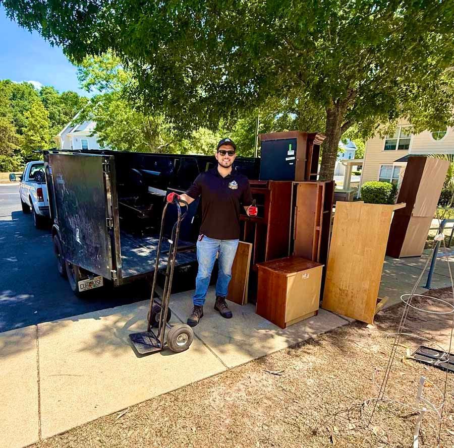 Person standing beside a dolly and a dumpster with furniture in a sunny outdoor area.