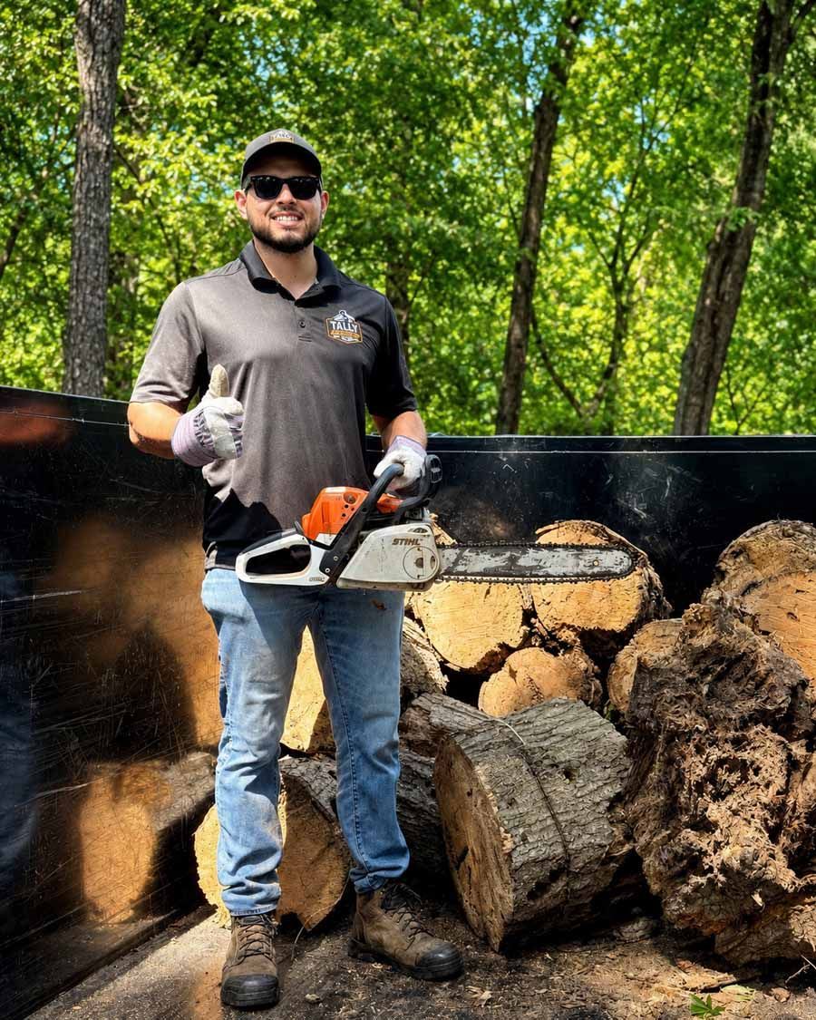 Man holding a chainsaw and log among stacked firewood outdoors in a wooded area