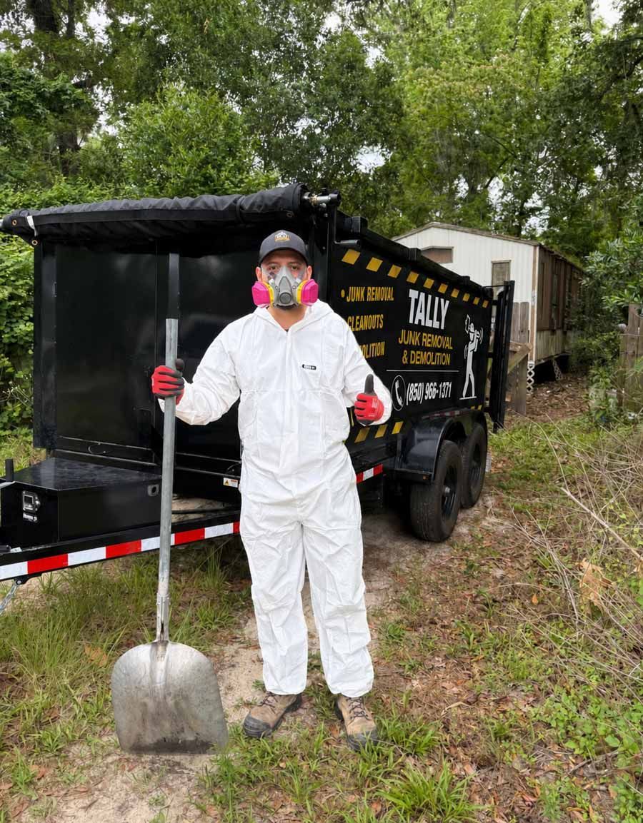 Person in white coveralls and red gloves holding a shovel beside a black trailer in a wooded area.