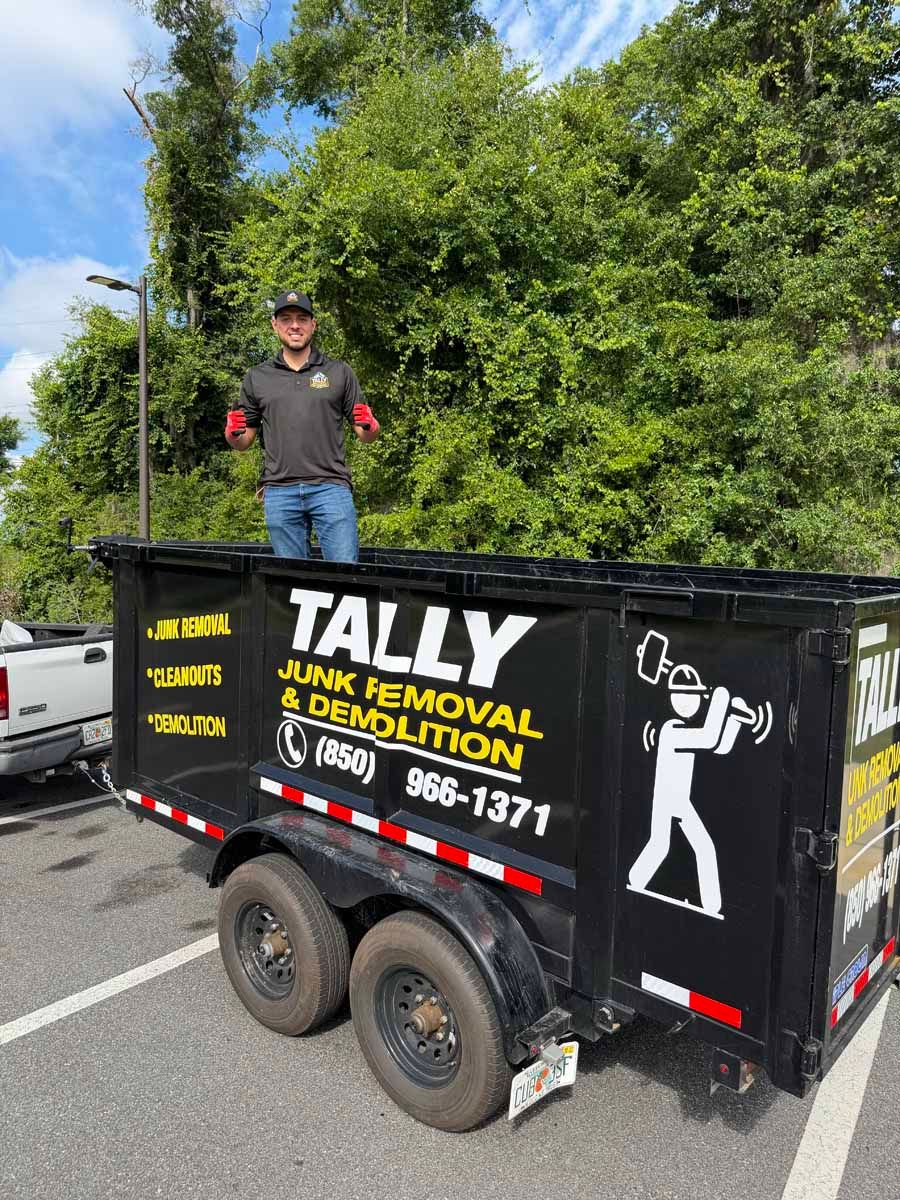 Worker standing in black dump trailer with Tally Junk Removal logo beside leafy roadside greenery
