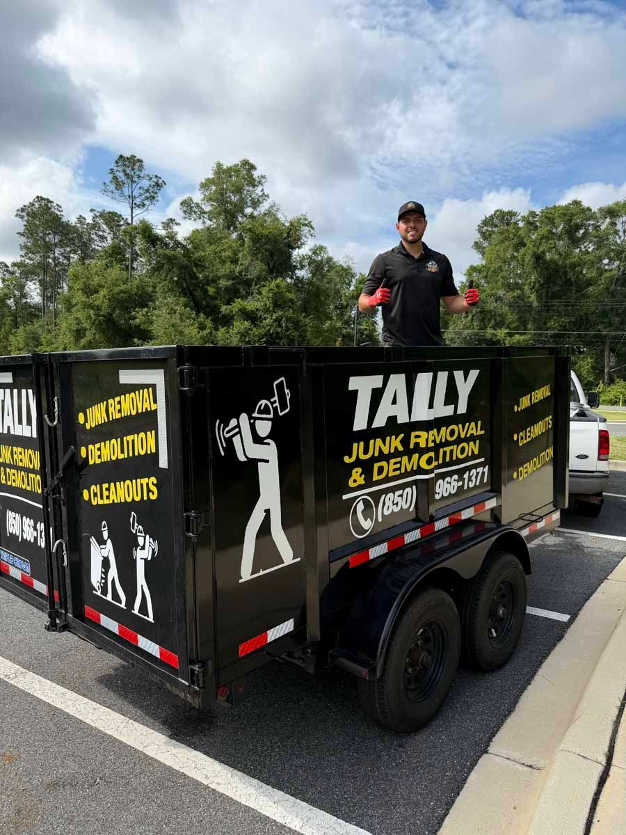 Man in a black truck bed with TALLY junk removal signage on a road under cloudy skies