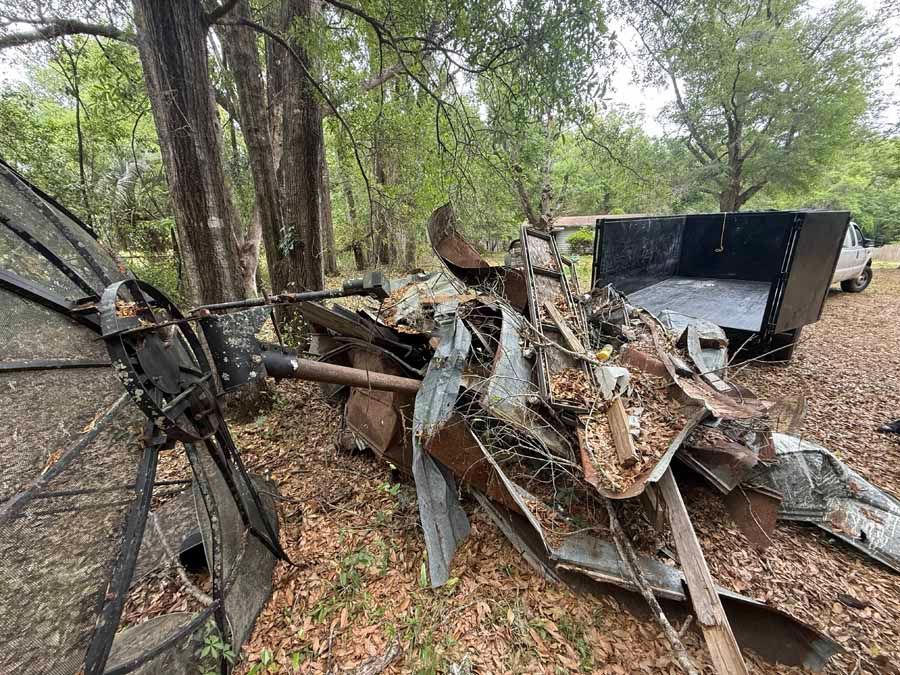 Twisted wreckage of a burned vehicle in a wooded area, with debris and blackened metal scattered around.