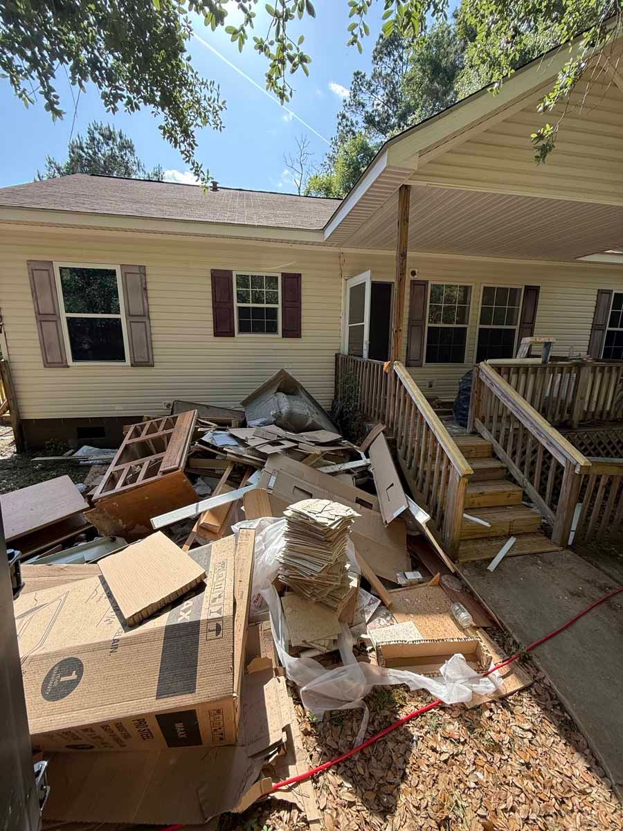 Rubble and discarded boxes piled beside a beige house with a covered porch and stairs.