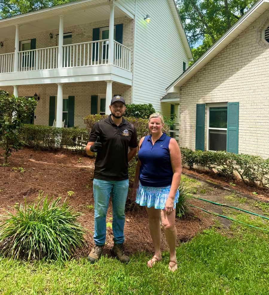 Two people standing in front of a house with white siding and green shutters, smiling outdoors.