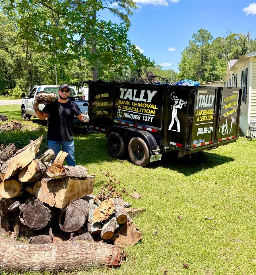 Man standing by a woodpile next to a black utility trailer in a grassy yard, with trees and a house in the background