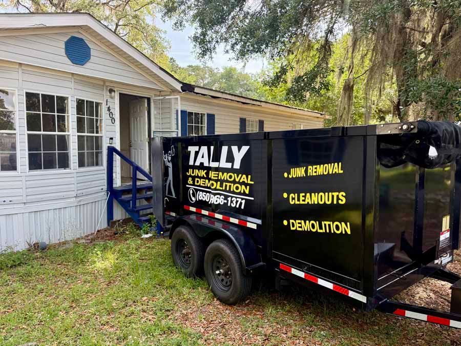 Black Tally demolition debris trailer parked beside a white house and blue steps