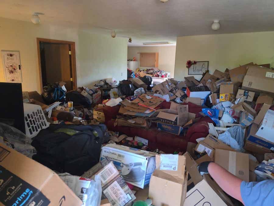 Cluttered living room packed with boxes, clothes, and belongings strewn across the floor and furniture.