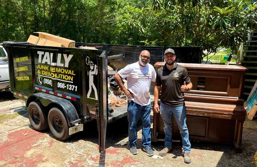 Two men stand beside a black trailer and a wood piano outdoors in a wooded yard.