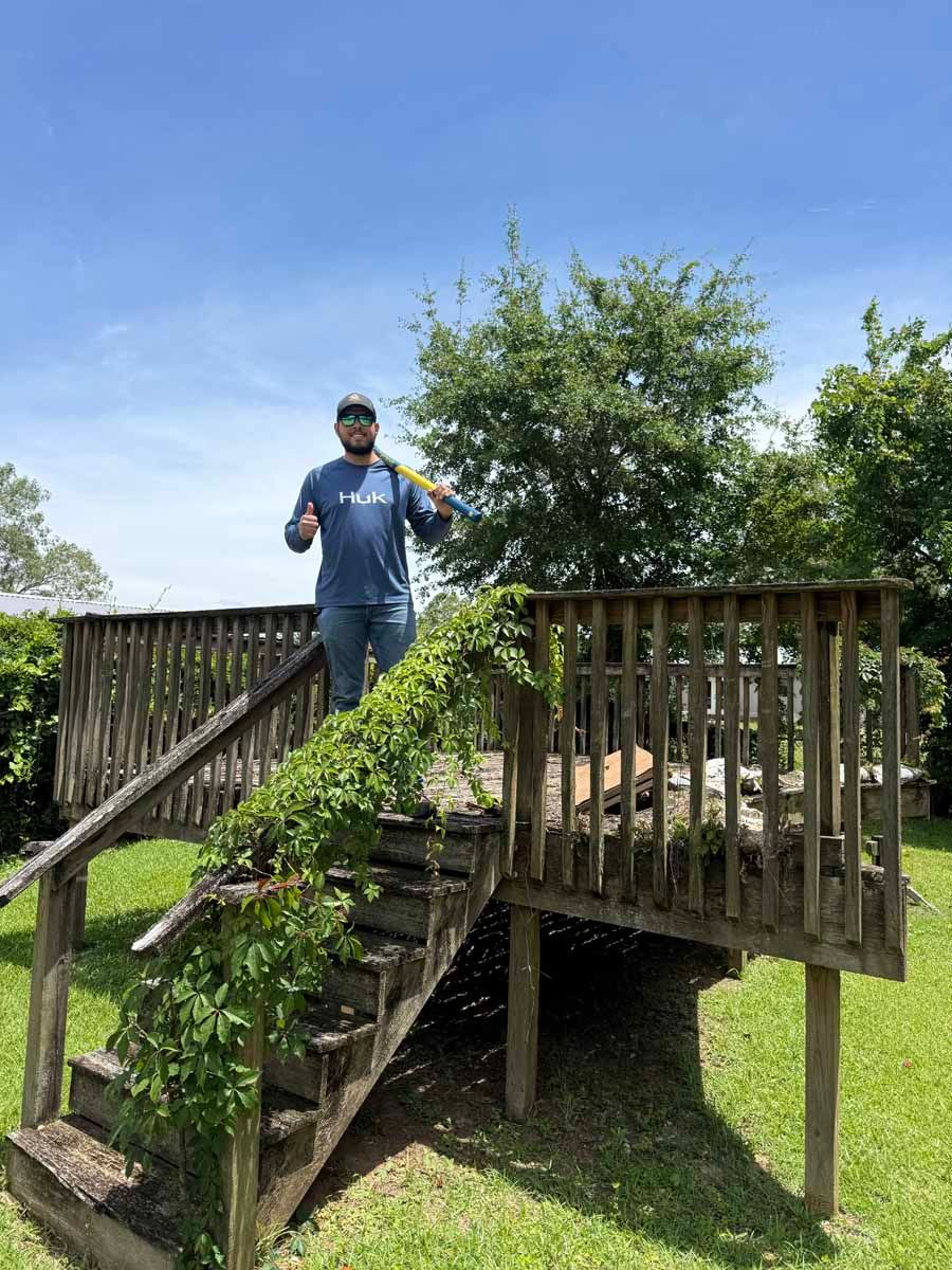 Person standing on a wooden staircase and deck outdoors, with a green tree and blue sky behind.