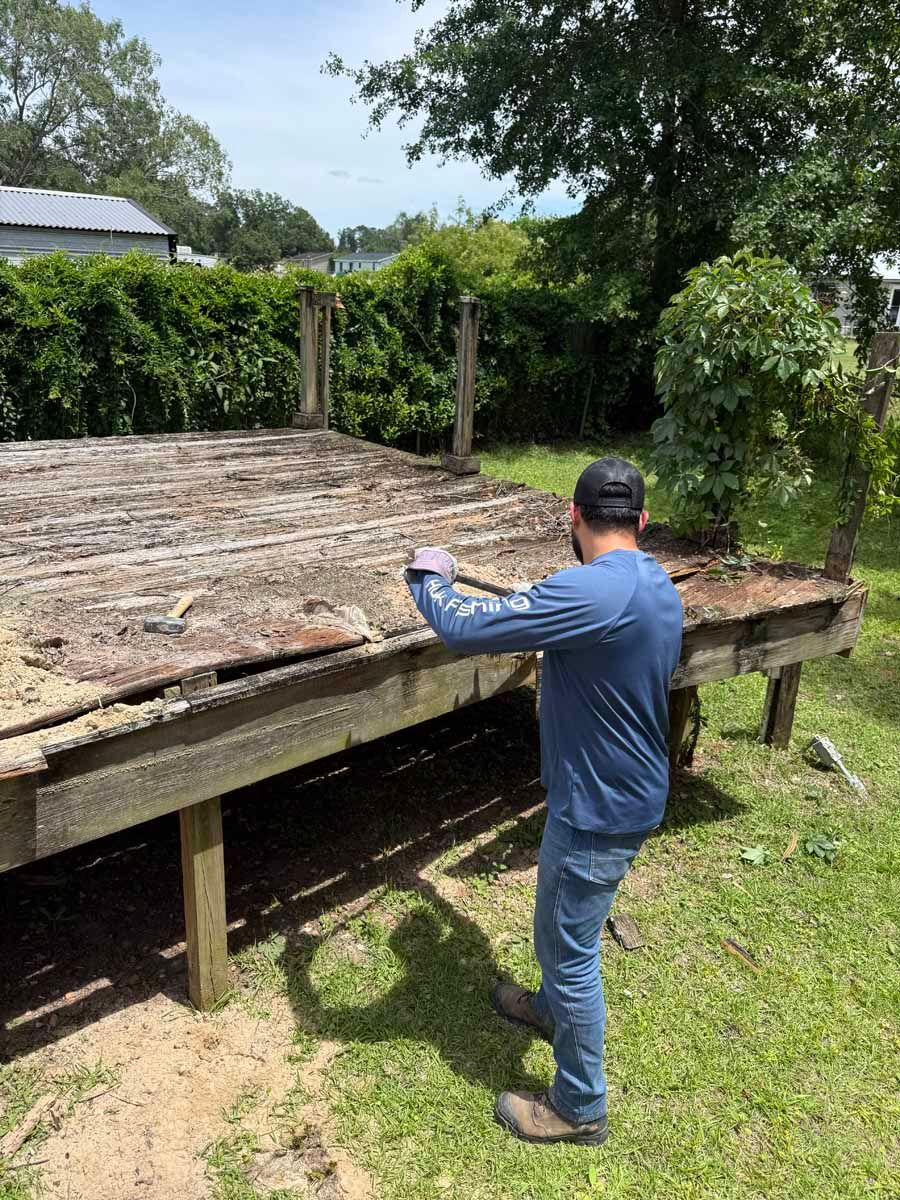 Person inspecting a raised wooden platform in a grassy outdoor yard with trees and hedges.