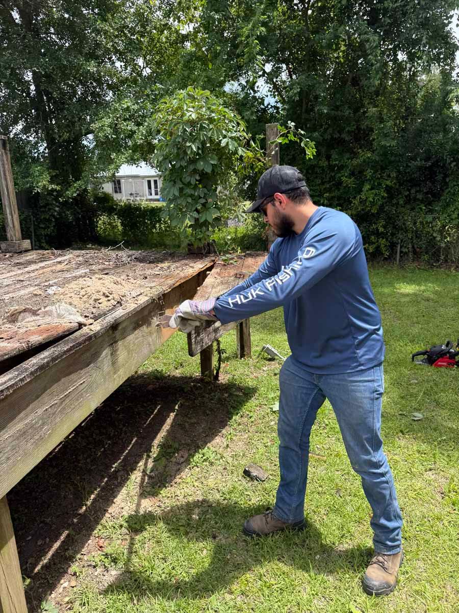 Man in blue shirt and cap pushing a large wooden beam in a grassy outdoor yard.