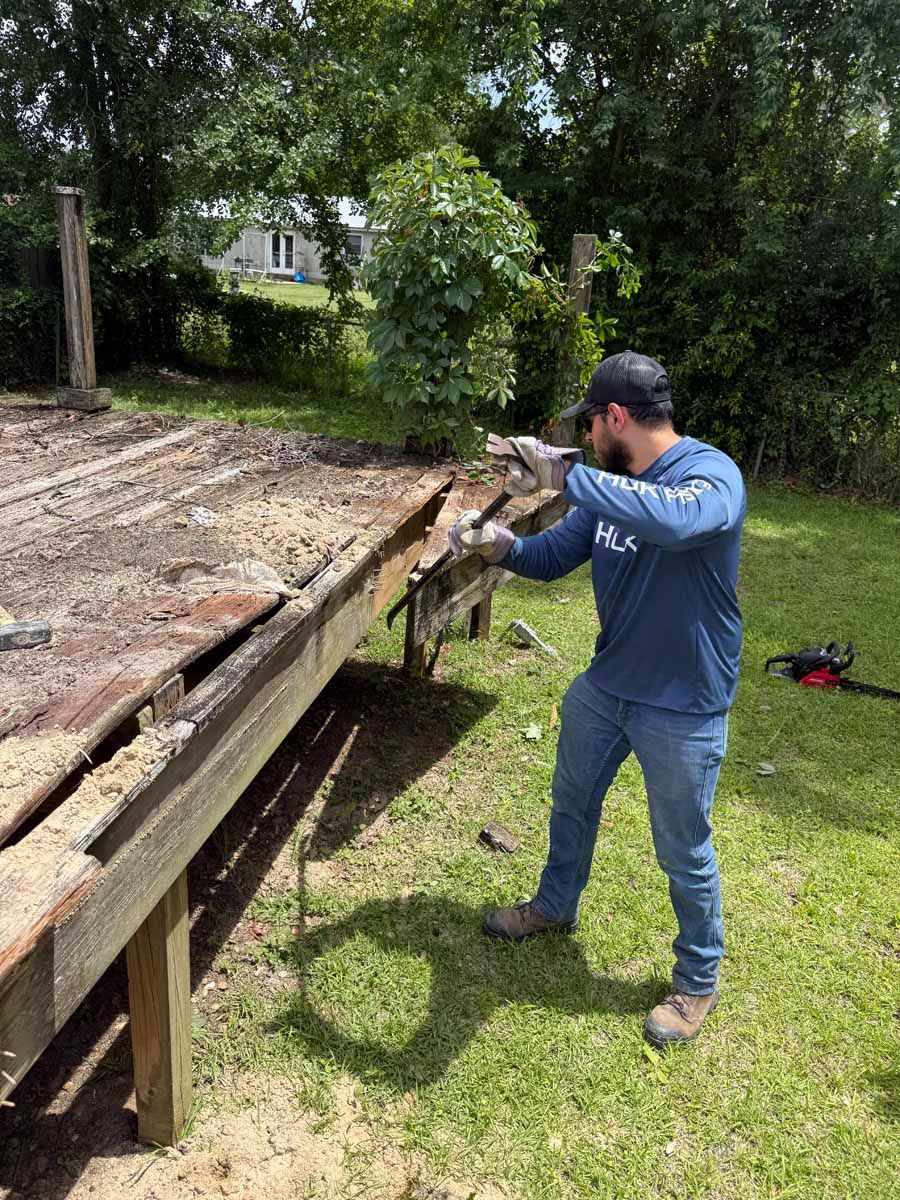 Man using a tool to pry up weathered wooden boards on a raised deck in a grassy yard